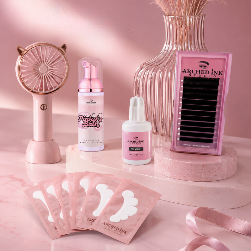 A pink beauty and lash care set arranged on a pink marble surface against a pink backdrop, including a handheld fan, two bottles labeled Arched Ink Studio, a box of Arched Ink Studio false eyelashes, and several pink adhesive patch packets. A ribbed glass vase with dried pampas grass sits behind the products.